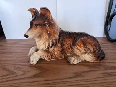 Ceramic collie pup statue, brown and cream, lying down on a wooden surface.